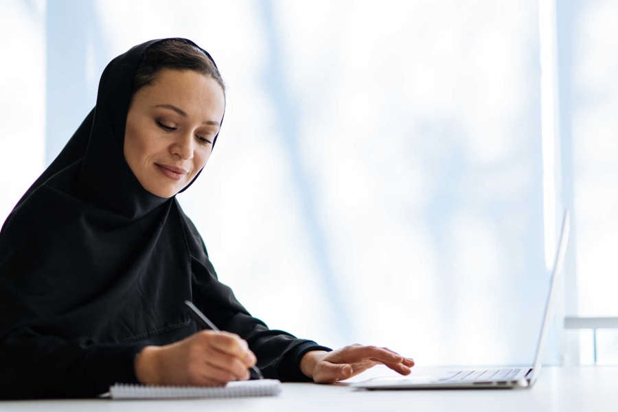 Emirati woman in black hijab and abaya writing in a notebook beside an open laptop at a sunlit desk