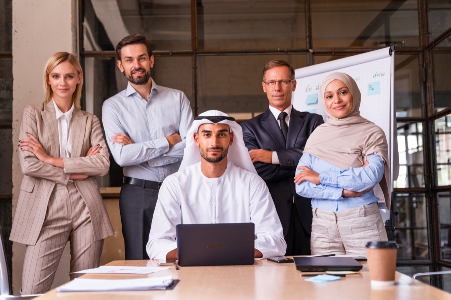 Diverse team portrait around an Emirati man with a laptop at a meeting table, with whiteboard behind
