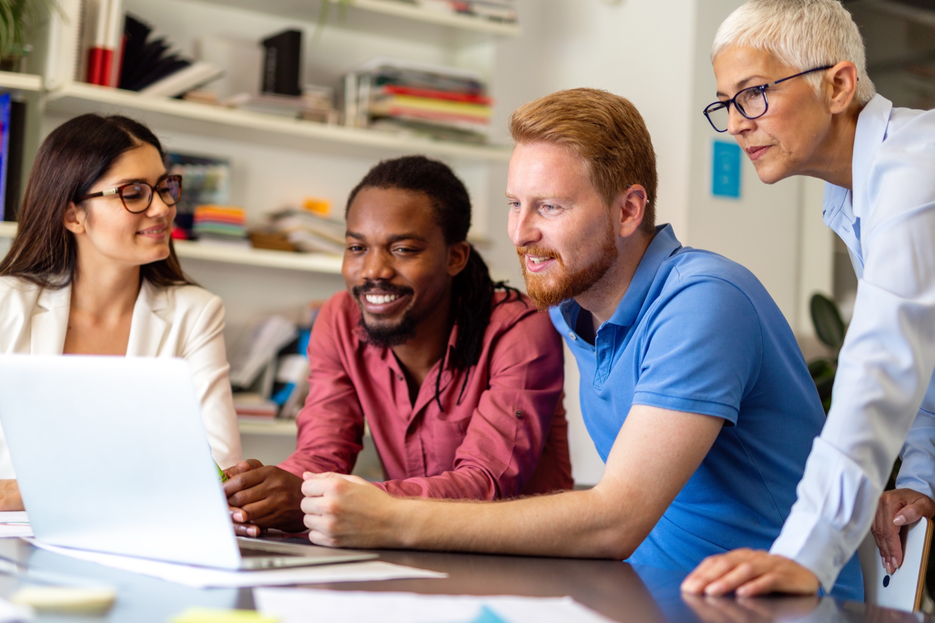Group of four diverse colleagues smiling as they look at a laptop screen together in an office