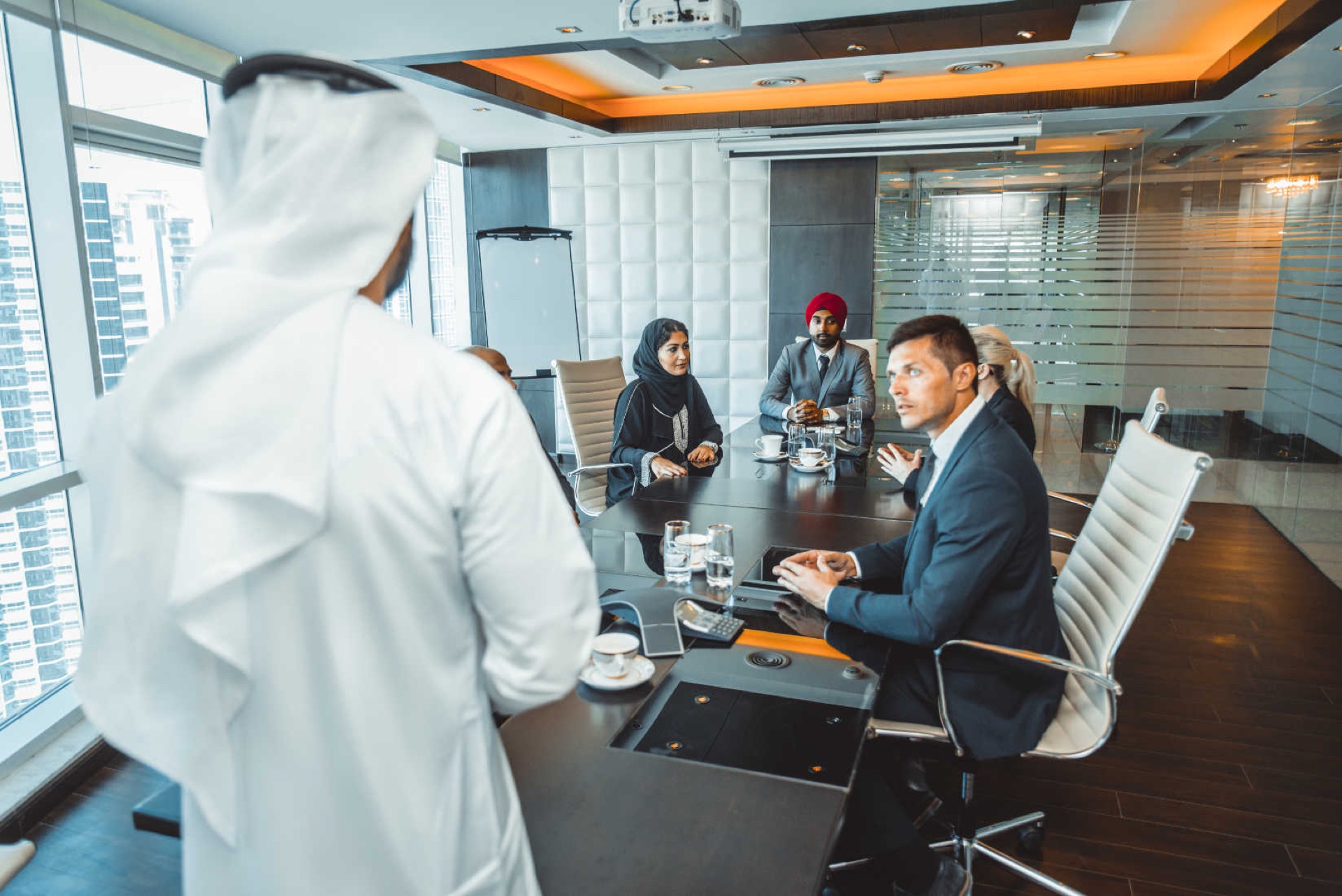 Emirati man in white kandura presenting to a diverse boardroom around a long meeting table in a high-rise office