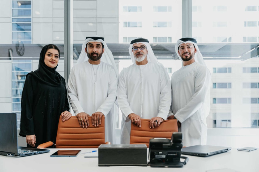 Four Emirati professionals in traditional dress standing behind a meeting table in front of a high-rise window