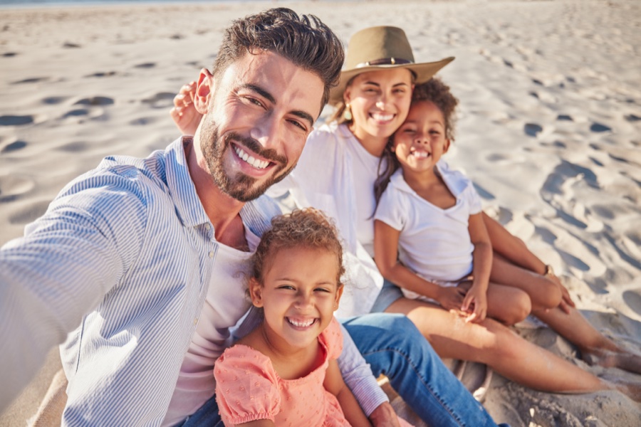 Family selfie of two adults and two young children sitting and smiling on a sandy beach